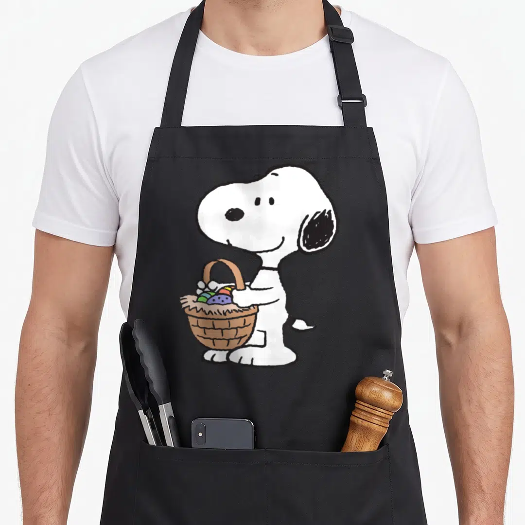 Funny easter basket apron - Snoopy standing holding brown basket with colorful eggs in cartoon style — shown on a black cotton canvas apron against a white background