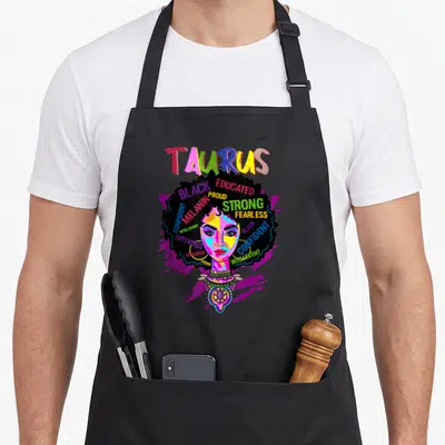 Afro april birthday apron - Cartoon black woman with TAURUS text and empowering words inside large afro hair on purple — shown on a black cotton canvas apron against a white background