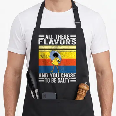 Chose these apron - Cartoon woman in yellow dress with blue umbrella against yellow orange blue stripes with white text — shown on a black cotton canvas apron against a white background