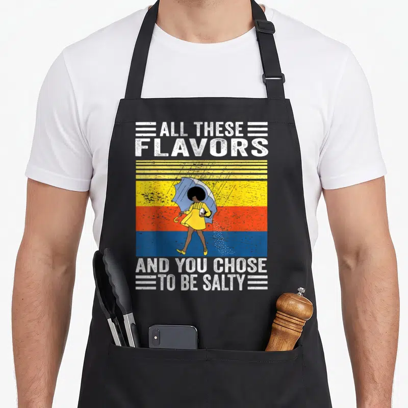 Chose these apron - Cartoon woman in yellow dress with blue umbrella against yellow orange blue stripes with white text — shown on a black cotton canvas apron against a white background