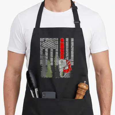 Thin wood apron - Vertical red chainsaw over grey distressed flag with green pine trees at bottom — shown on a black cotton canvas apron against a white background
