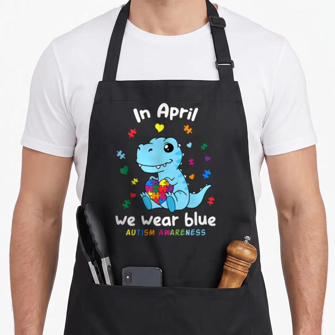 Baby april apron - Blue cartoon dinosaur holding rainbow puzzle heart surrounded by colorful puzzle pieces and hearts — shown on a black cotton canvas apron against a white background