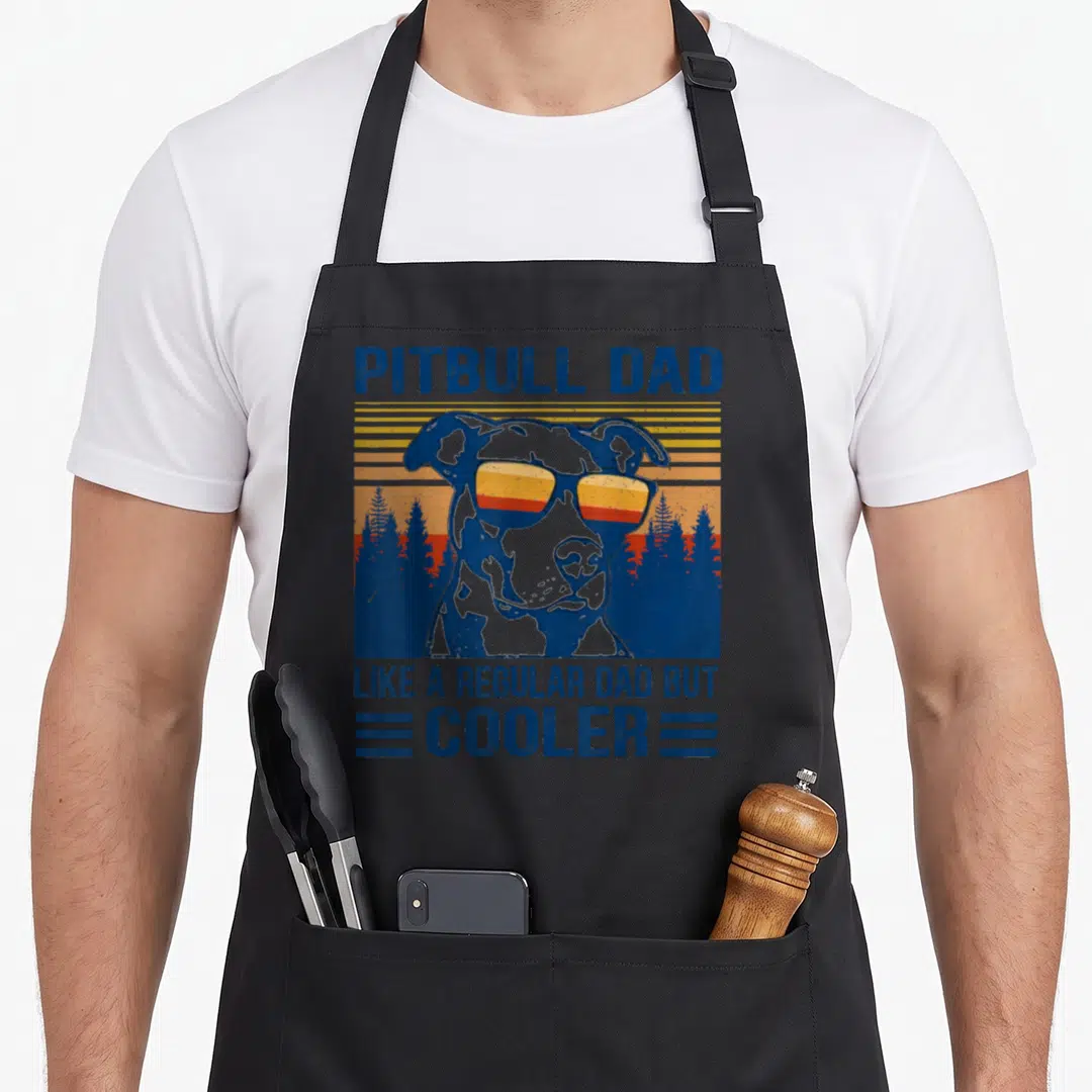 Pitbull dad but apron - Retro pitbull wearing sunglasses against striped sunset with pine trees and blue text — shown on a black cotton canvas apron against a white background