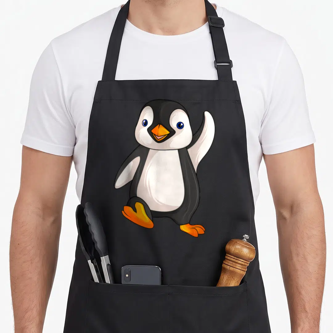 Penguin aprons for grilling - Cartoon penguin waving one flipper with orange beak and feet on black background — shown on a black cotton canvas apron against a white background