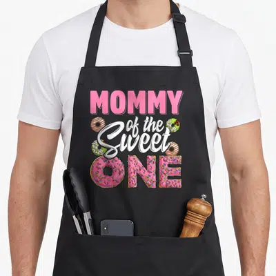 Sweet birthday donut apron - MOMMY of the Sweet ONE text with pink sprinkled and brown glazed donuts — shown on a black cotton canvas apron against a white background