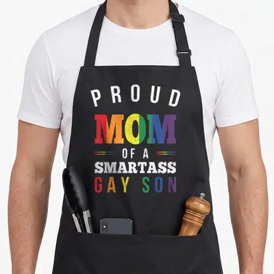 Smartass gay pride apron - Stacked rainbow text reading PROUD MOM OF A SMARTASS GAY SON with distressed effect — shown on a black cotton canvas apron against a white background