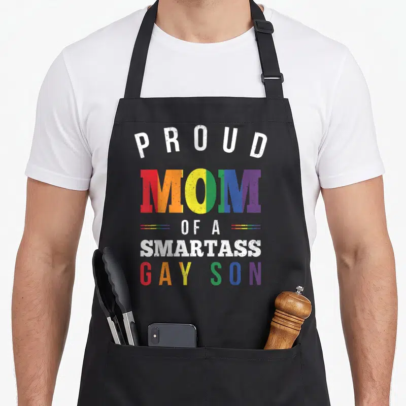 Smartass gay pride apron - Stacked rainbow text reading PROUD MOM OF A SMARTASS GAY SON with distressed effect — shown on a black cotton canvas apron against a white background