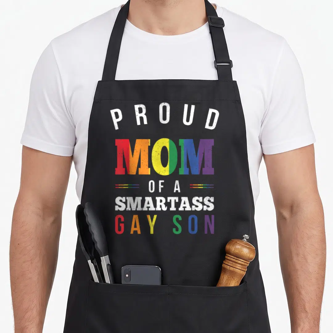 Smartass gay pride apron - Stacked rainbow text reading PROUD MOM OF A SMARTASS GAY SON with distressed effect — shown on a black cotton canvas apron against a white background