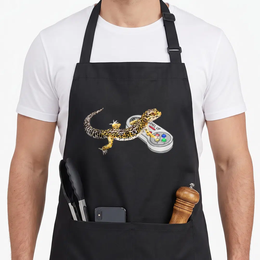 Leopard video game apron - Realistic yellow spotted leopard gecko draped over a grey game controller with LEVEL UP text — shown on a black cotton canvas apron against a white background