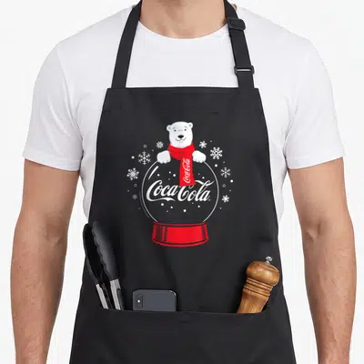 Snowglobe bear apron - Polar bear in red scarf atop snow globe with Coca-Cola logo and white snowflakes — shown on a black cotton canvas apron against a white background