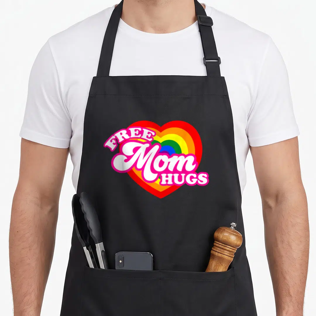 Free hug gay pride apron - Rainbow heart with arched FREE Mom HUGS text in white and pink cartoon lettering — shown on a black cotton canvas apron against a white background