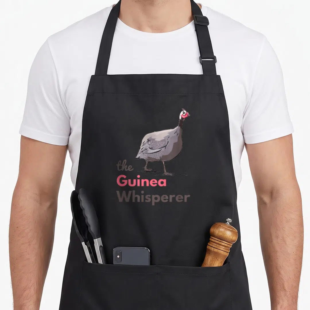 Guineafowl hen apron - Cartoon guinea fowl standing above stacked text reading the Guinea Whisperer in grey pink — shown on a black cotton canvas apron against a white background