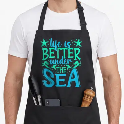 Vacation mermaid apron - Stacked blue and teal text life is better under the sea with starfish and mermaid silhouettes — shown on a black cotton canvas apron against a white background