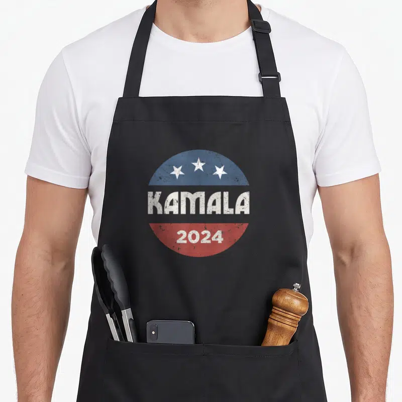 Kamala presidents apron - Circular 2024 campaign logo with three stars and KAMALA text across blue white red bands — shown on a black cotton canvas apron against a white background