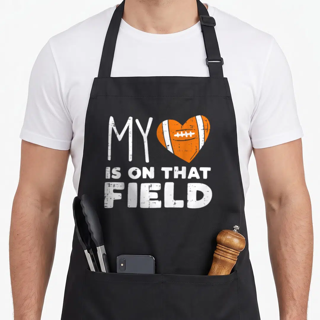 Field football apron - Distressed white text MY next to an orange heart shaped like a football with laces — shown on a black cotton canvas apron against a white background