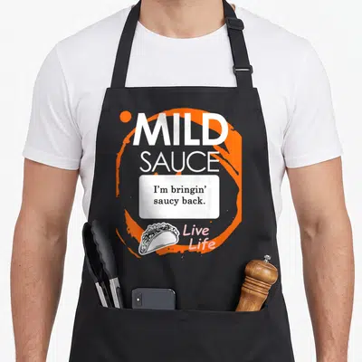 Taco sauce halloween apron - Mild sauce text with I'm bringin saucy back and taco illustration on orange splash — shown on a black cotton canvas apron against a white background