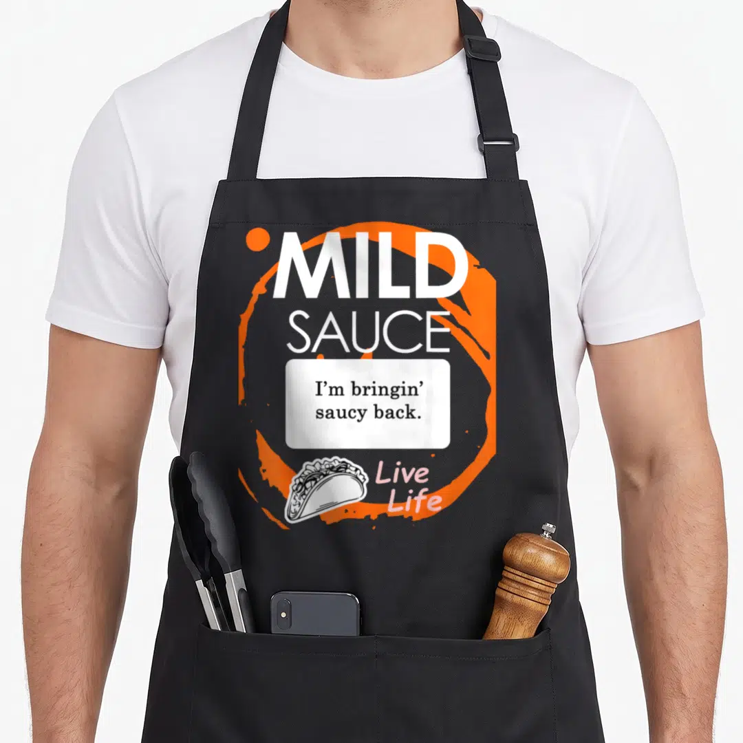 Taco sauce halloween apron - Mild sauce text with I'm bringin saucy back and taco illustration on orange splash — shown on a black cotton canvas apron against a white background