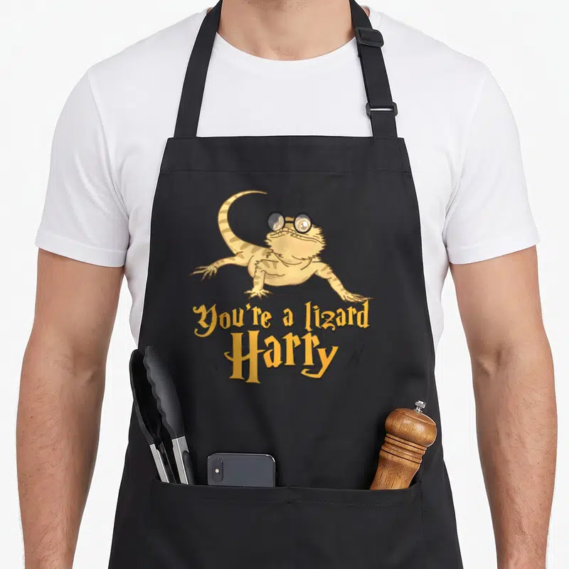 Harry lizard apron - Bearded dragon with glasses and You're a lizard Harry text flanked by lightning bolts — shown on a black cotton canvas apron against a white background