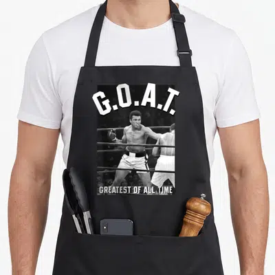 Muhammad sports legendz apron - Boxer in ring with arched G.O.A.T. text and straight Greatest of All Time caption below — shown on a black cotton canvas apron against a white background