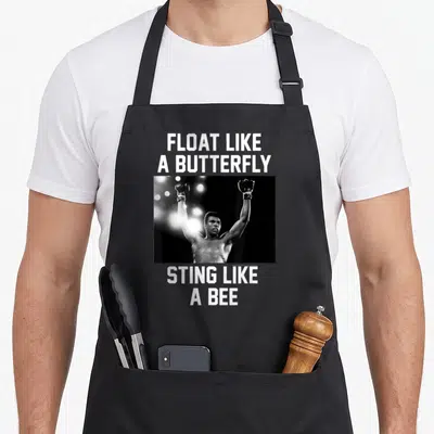 Muhammad butterfly apron - Grayscale photo of boxer between white text reading float like a butterfly sting like a bee — shown on a black cotton canvas apron against a white background