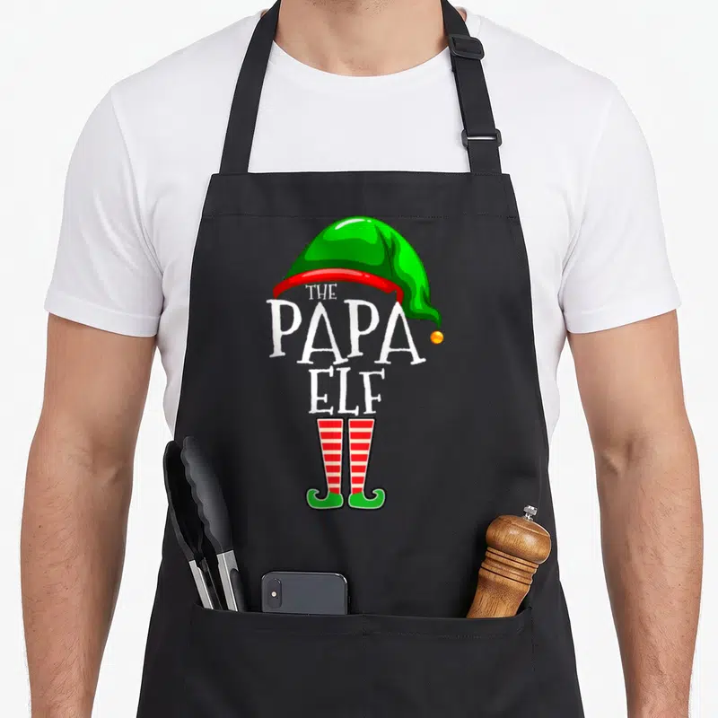Papa christmas elf apron - Stacked THE PAPA ELF text with green hat and red striped elf shoes — shown on a black cotton canvas apron against a white background