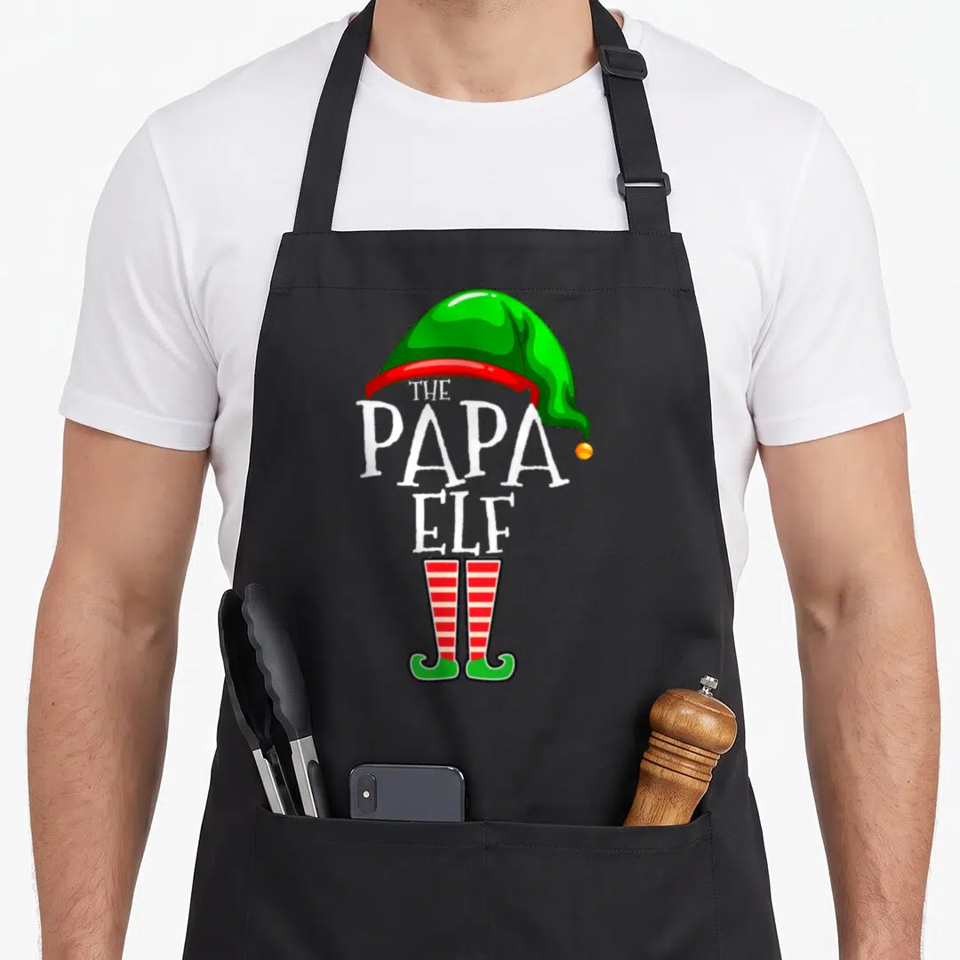 Papa christmas elf apron - Stacked THE PAPA ELF text with green hat and red striped elf shoes — shown on a black cotton canvas apron against a white background