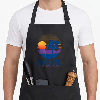 Tree artistic apron - Retro circular graphic with Joshua tree, striped sun, purple mountains, and text in blue — shown on a black cotton canvas apron against a white background