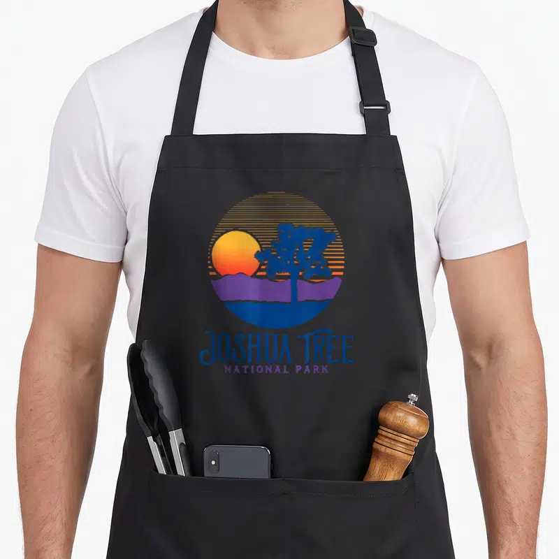 Tree artistic apron - Retro circular graphic with Joshua tree, striped sun, purple mountains, and text in blue — shown on a black cotton canvas apron against a white background