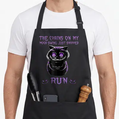 Chain snapped aprons - Black cartoon cat with purple eyes and text THE CHAINS ON MY MOOD SWING JUST SNAPPED RUN between — shown on a black cotton canvas apron against a white background