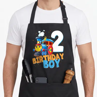 Birthday train 2nd apron - Cartoon blue train with number 2 and stacked BIRTHDAY BOY text in orange and blue — shown on a black cotton canvas apron against a white background