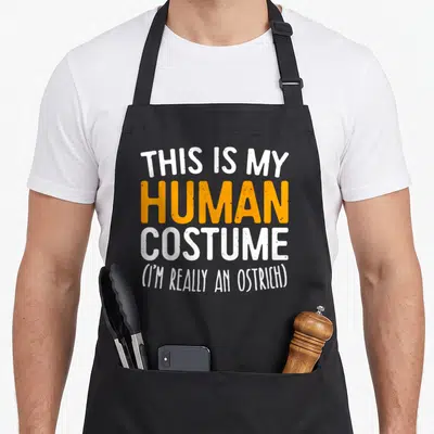 Human costume i love my ostrich apron - Stacked white and orange text reading THIS IS MY HUMAN COSTUME I'M REALLY — shown on a black cotton canvas apron against a white background