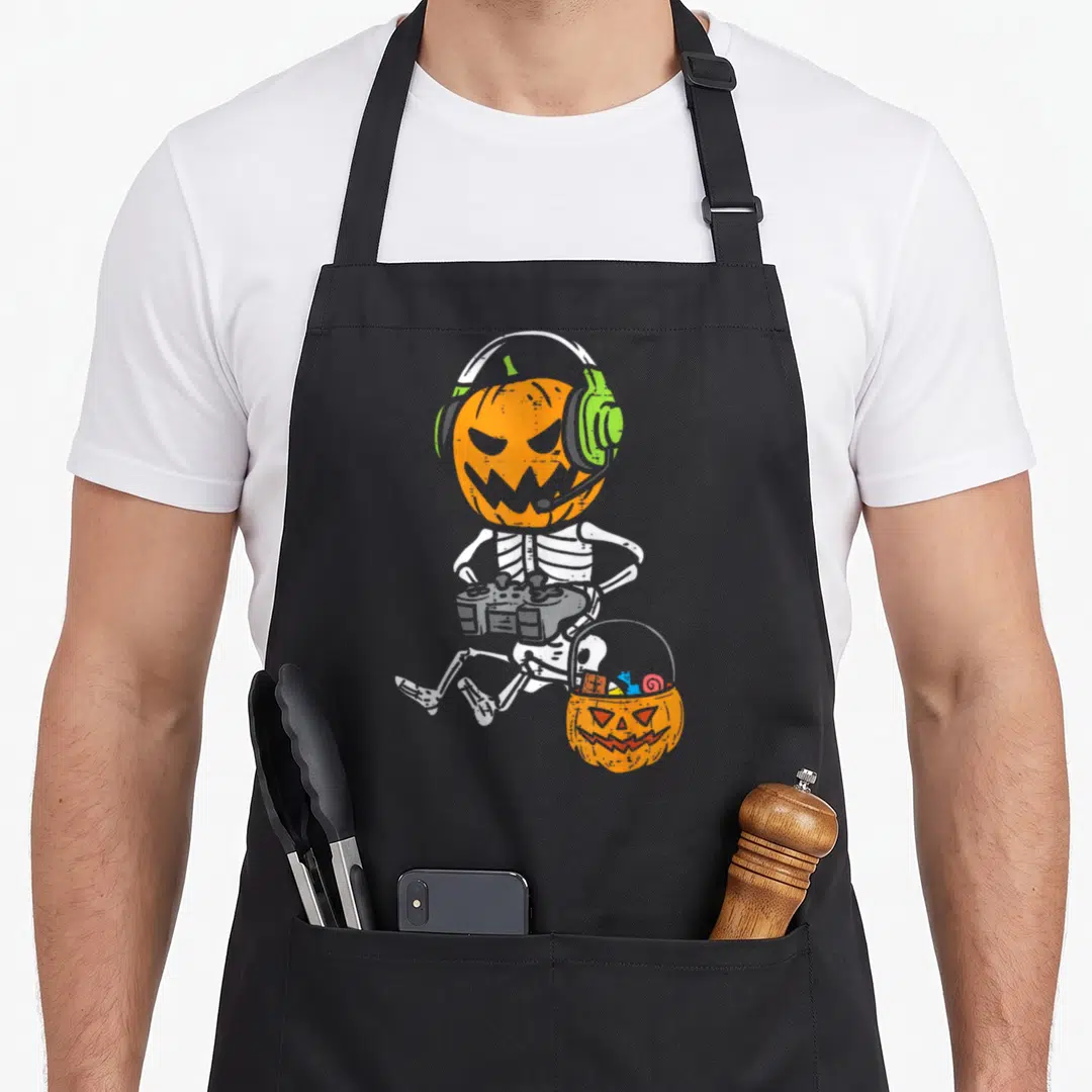 Gamer pumpkin skeleton halloween apron - Cartoon skeleton with pumpkin head, green headphones, grey controller — shown on a black cotton canvas apron against a white background