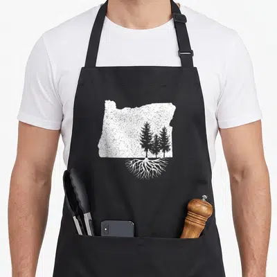 Root oregon apron - White Oregon state silhouette with three pine trees and roots in distressed style — shown on a black cotton canvas apron against a white background