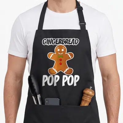 Pop christmas gingerbread apron - Cartoon gingerbread man with green bowtie between GINGERBREAD and POP POP text — shown on a black cotton canvas apron against a white background