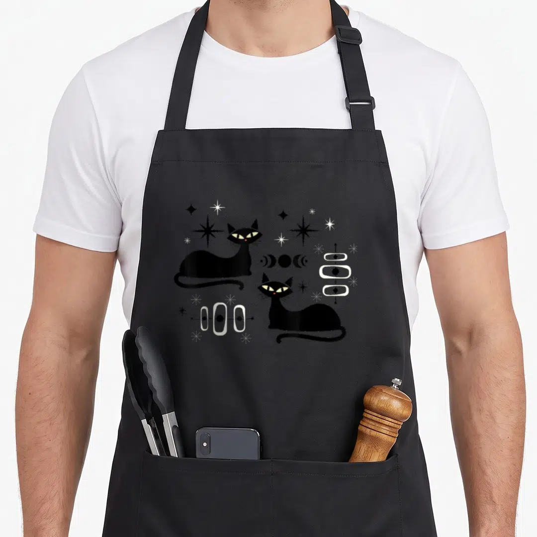 Atomic modern apron - Two black cats face diagonally amid stars, moon phases, and atomic symbols in retro style — shown on a black cotton canvas apron against a white background
