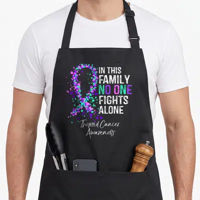 Thyroid family aprons - Heart ribbon in teal purple pink white next to In This Family No One Fights Alone text — shown on a black cotton canvas apron against a white background