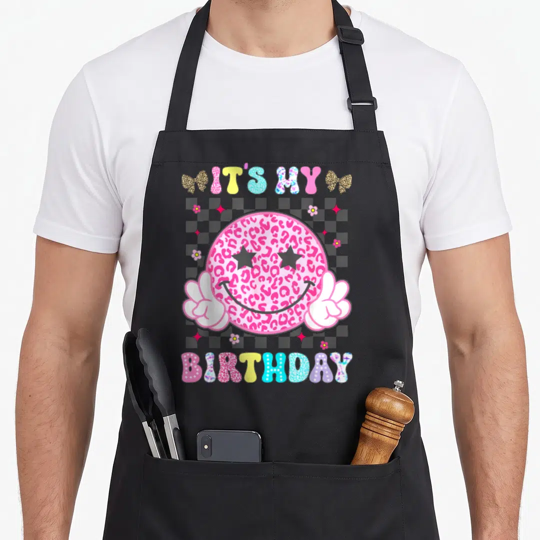 Smile custom face apron - Pink leopard smiley face with star eyes and peace hands saying IT'S MY BIRTHDAY on checkered — shown on a black cotton canvas apron against a white background