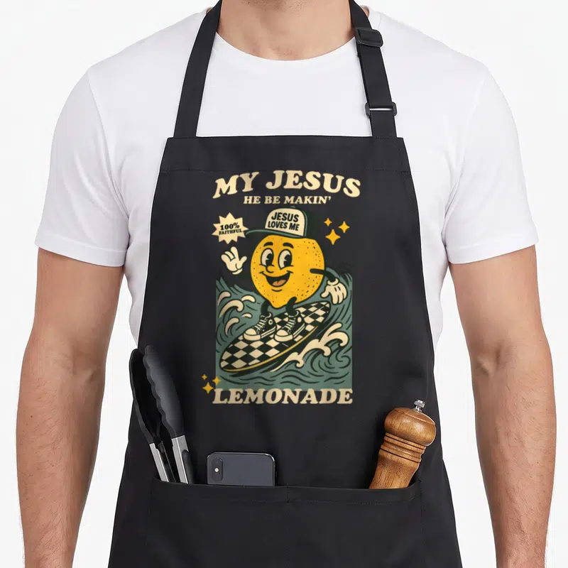 Making lemonade apron - Cartoon lemon surfing checkerboard board with MY JESUS HE BE MAKIN LEMONADE text — shown on a black cotton canvas apron against a white background