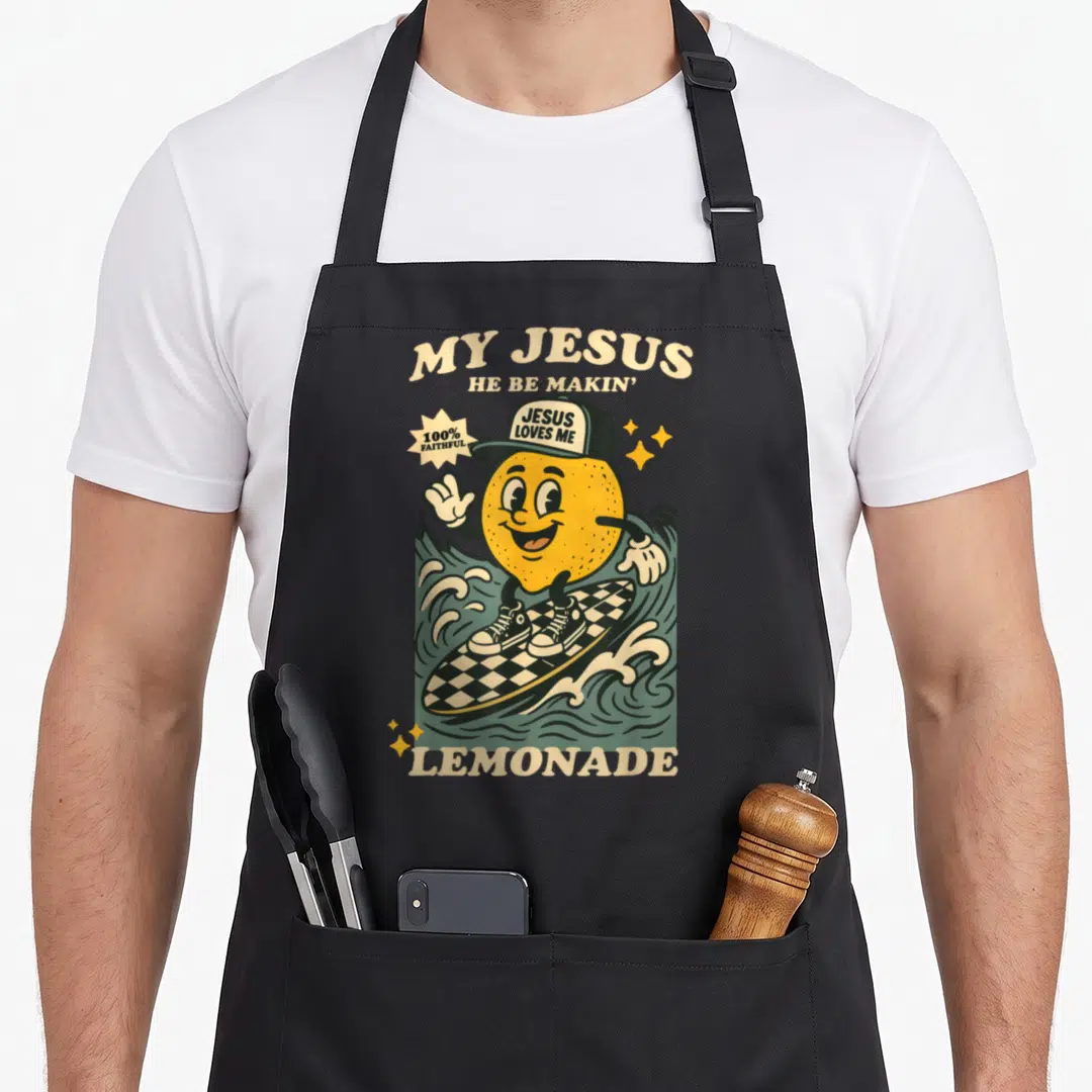 Making lemonade apron - Cartoon lemon surfing checkerboard board with MY JESUS HE BE MAKIN LEMONADE text — shown on a black cotton canvas apron against a white background
