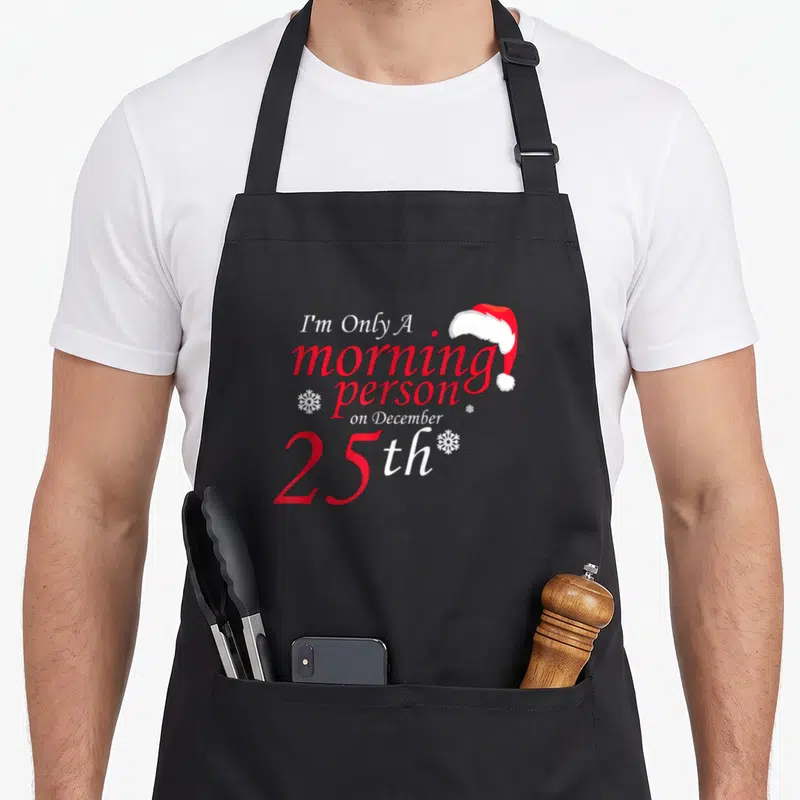 25th women's christmas apron - Red and white text I'm Only A morning person on December 25th with Santa hat — shown on a black cotton canvas apron against a white background