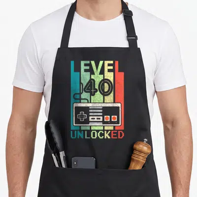 Level 40th apron - Retro game controller on teal green yellow red stripes with LEVEL 40 UNLOCKED text — shown on a black cotton canvas apron against a white background