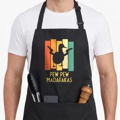 Pistol cotton duck aprons - Grey duck silhouette holding two guns over orange, yellow, teal, and grey stripes with text — shown on a black cotton canvas apron against a white background