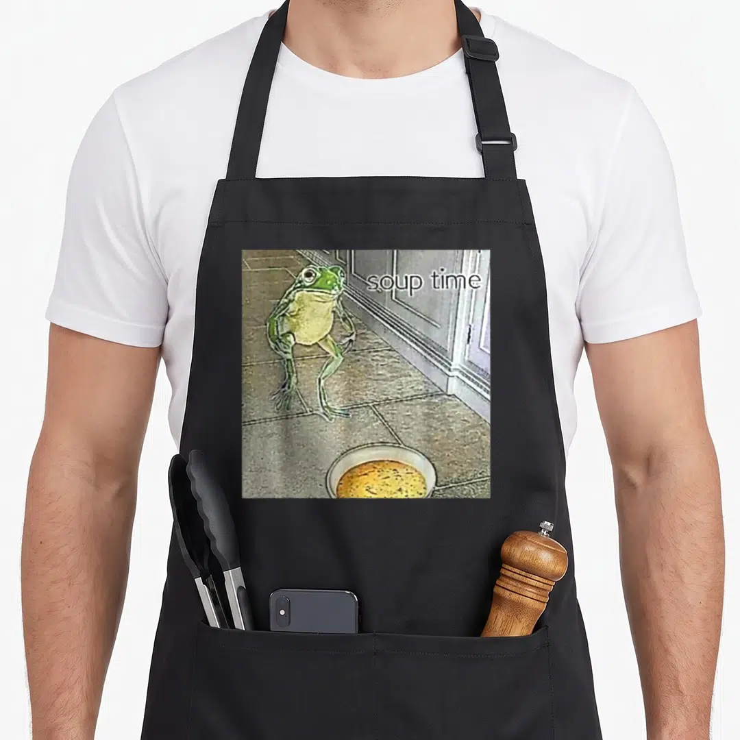Oddly meme apron - Frog standing on grey tiles near yellow soup bowl with text soup time — shown on a black cotton canvas apron against a white background