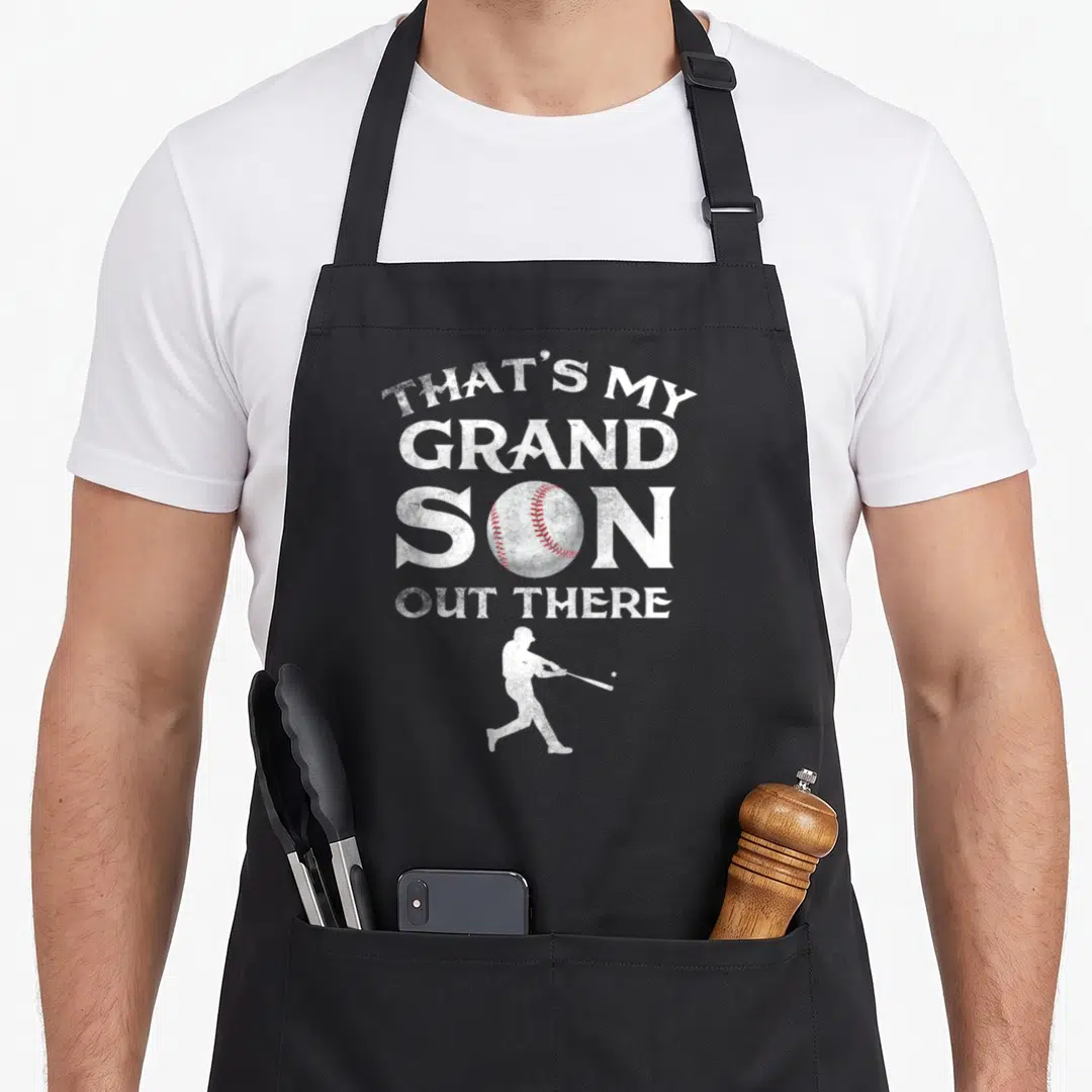 Grandson baseball apron - Distressed white text reading That's My Grand Son Out There with red stitched baseball — shown on a black cotton canvas apron against a white background