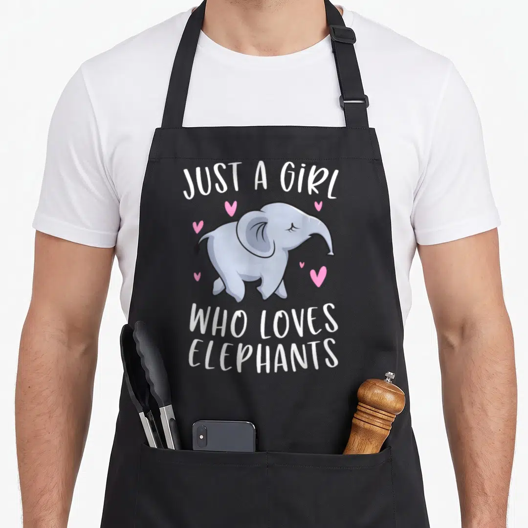 Vintage elephant print apron - Cartoon blue-grey elephant with five pink hearts and white text reading JUST A GIRL WHO — shown on a black cotton canvas apron against a white background