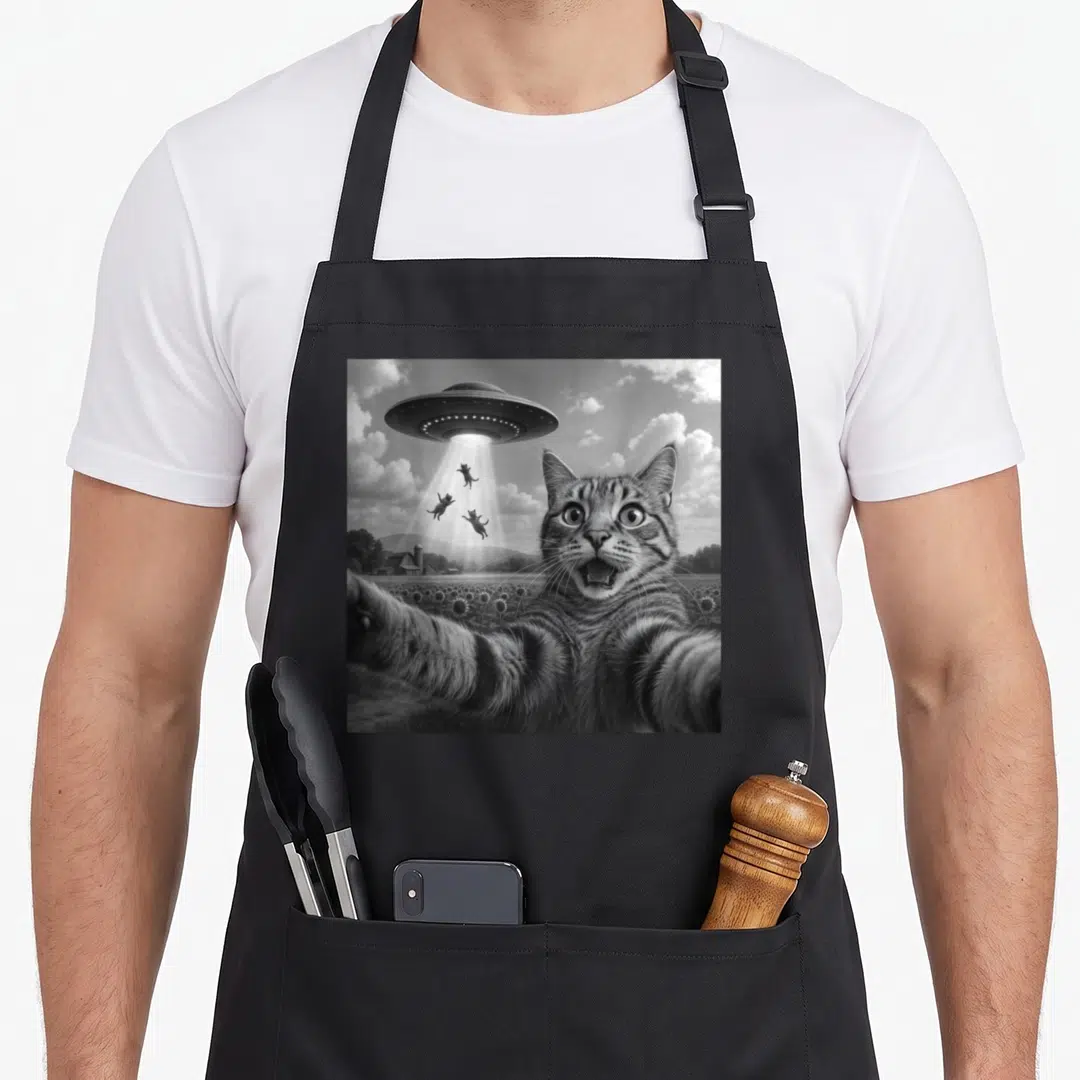 Alien funny cat apron - Black and white cat selfie with UFO abducting three cats over sunflowers — shown on a black cotton canvas apron against a white background