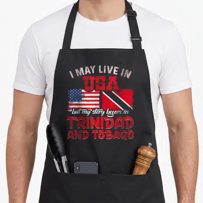 Trinidad story of the apron - Two flags and red white text reading I MAY LIVE IN USA but my story began in TRINIDAD — shown on a black cotton canvas apron against a white background