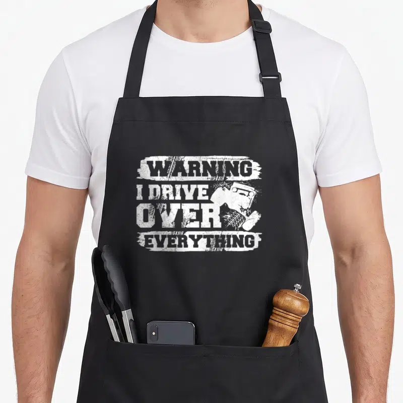 Crawling crawler apron - White stacked text WARNING I DRIVE OVER EVERYTHING with off-road vehicle and tire tracks — shown on a black cotton canvas apron against a white background