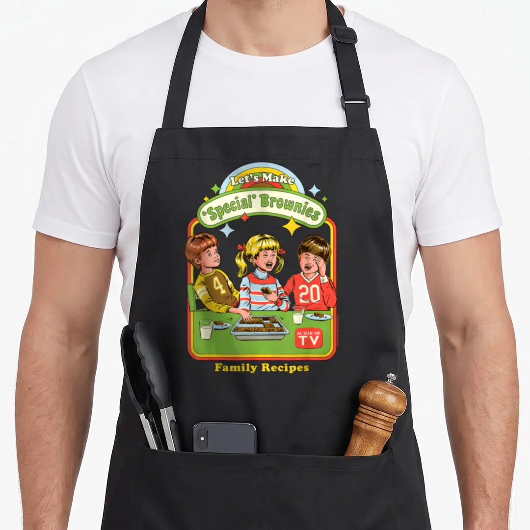 Browny special aprons - Three children eating brownies at green table under rainbow and Let's Make Special Brownies text — shown on a black cotton canvas apron against a white background