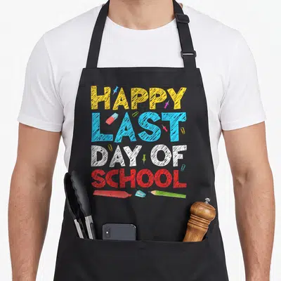 Funny school teacher apron - HAPPY LAST DAY OF SCHOOL text with eraser sharpener pencil and push pins — shown on a black cotton canvas apron against a white background